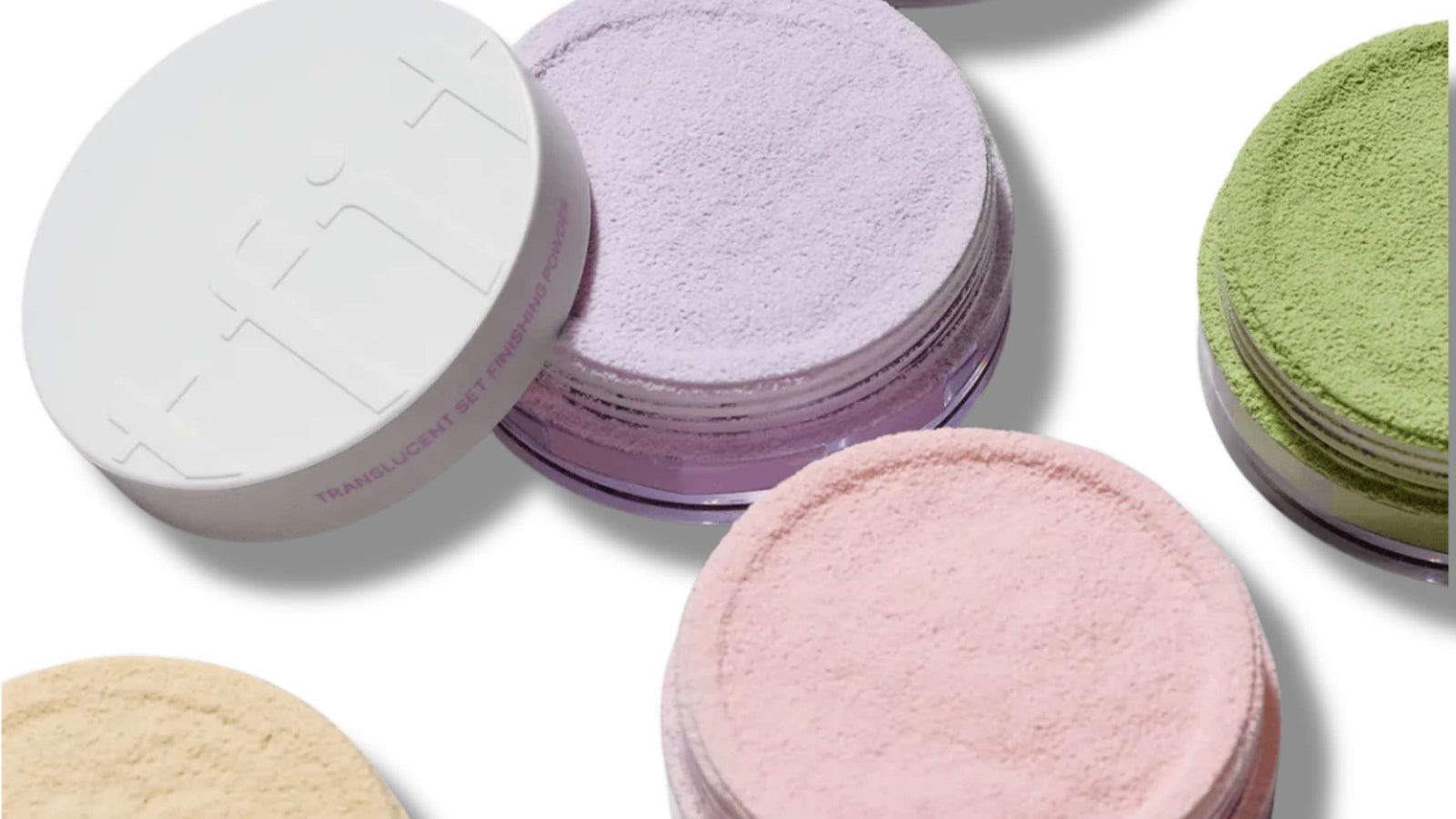 Närbild på olika nyanser av lösa, genomskinliga setting-puder i behållare. Pudren visas i lavendel, rosa och gröna nyanser, där lavendelpudret är i förgrunden.