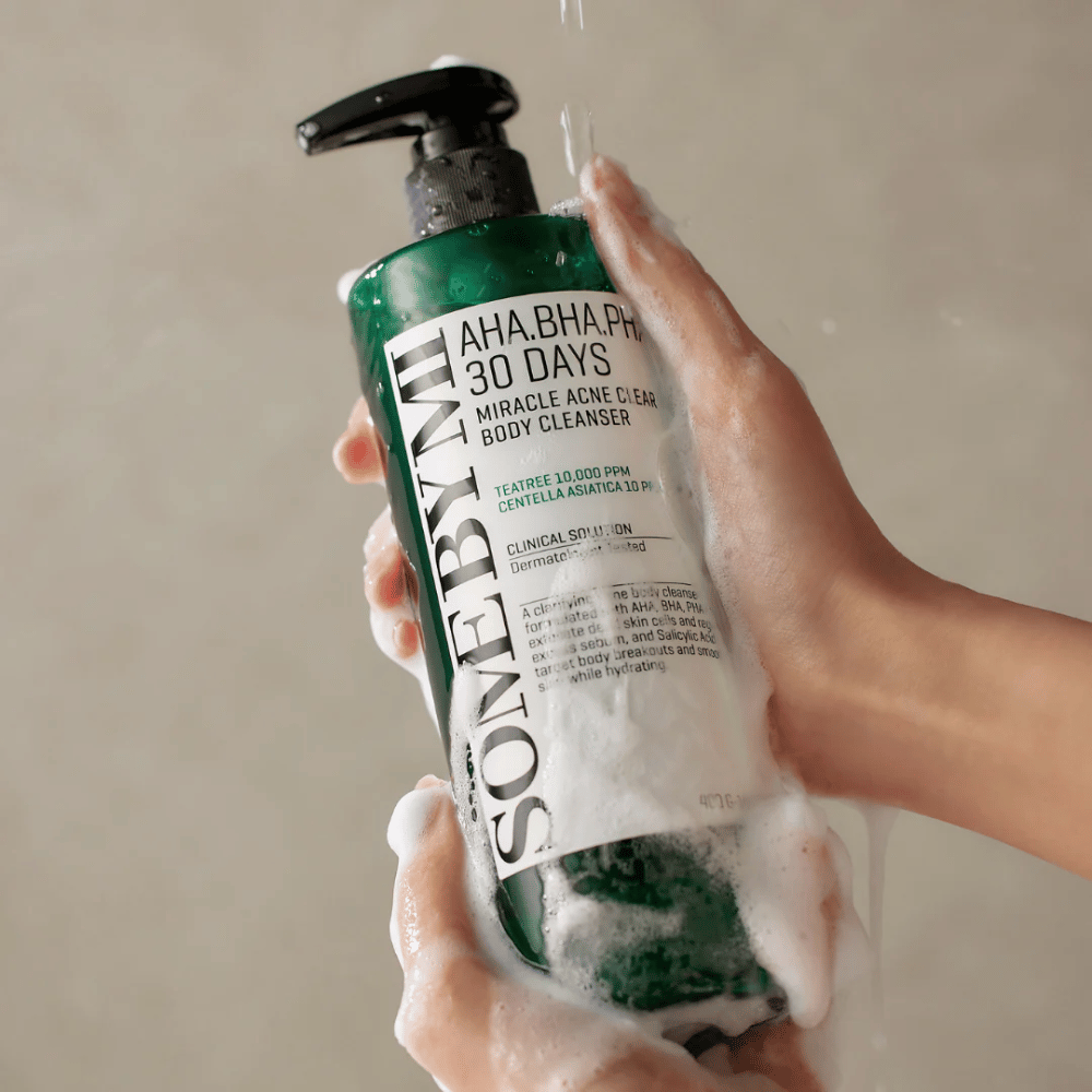 Une personne tient un flacon SOME BY MI AHA.BHA.PHA 30 Days Miracle Acne Clear Body Cleanser. Le flacon est vert avec une étiquette blanche et est couvert de mousse du nettoyant. La personne se lave avec le produit tandis que l'eau coule sur ses mains.