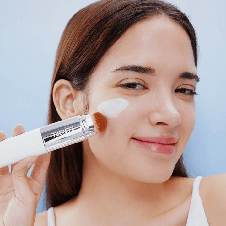 Un modèle applique ELROEL Blanc Cover Cream Stick concealer sur la joue avec un pinceau intégré pour une couverture naturelle et uniforme.