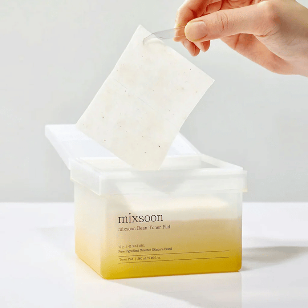 Une main tient une pince et soulève un Mixsoon Bean Toner Pad d'un récipient carré et transparent avec une teinte jaunâtre. Le récipient contient 70 pads de toner et a un texte décrivant le produit.