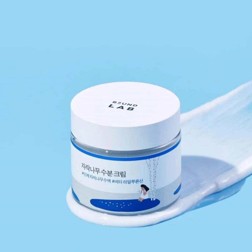 Round Lab Birch Juice Moisturizing Cream dans un pot teinté de bleu avec couvercle blanc, debout dans une couche de crème sur fond bleu.