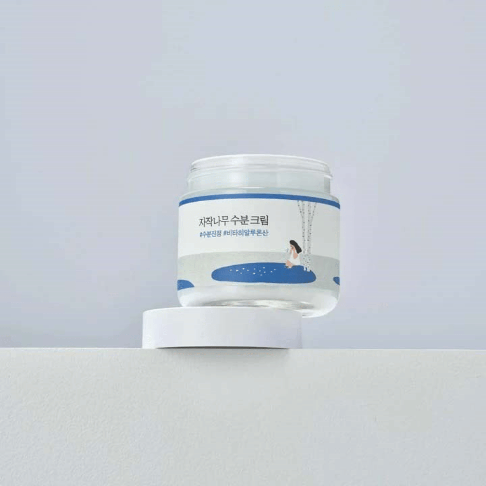Round Lab Birch Juice Moisturizing Cream dans un pot transparent avec étiquette blanche et illustration bleue d'une femme près d'un bouleau, debout sur une surface blanche contre un fond clair.