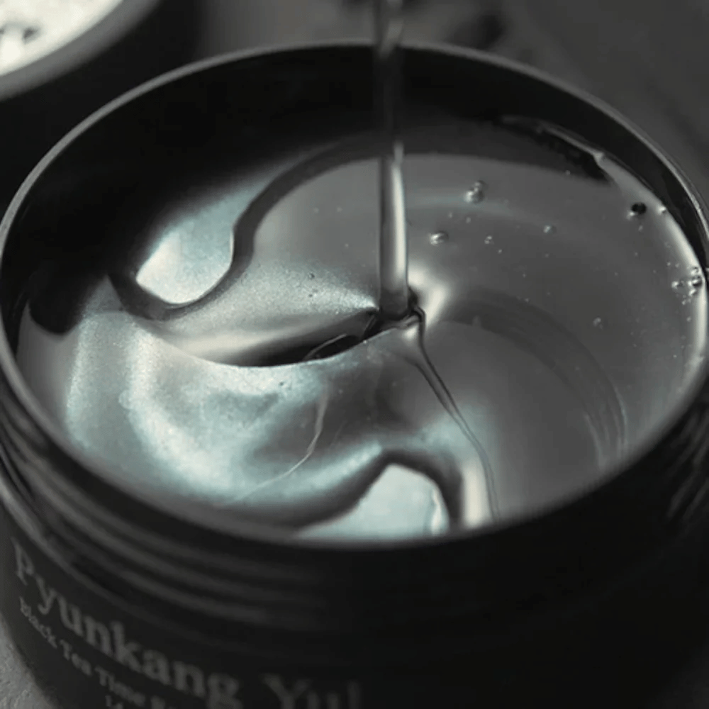 Gros plan sur des patchs contour des yeux en hydrogel noirs dans un pot ouvert de Pyunkang Yul. Les patchs brillants baignent dans le sérum et mettent en valeur le caractère hydratant et luxueux du produit.