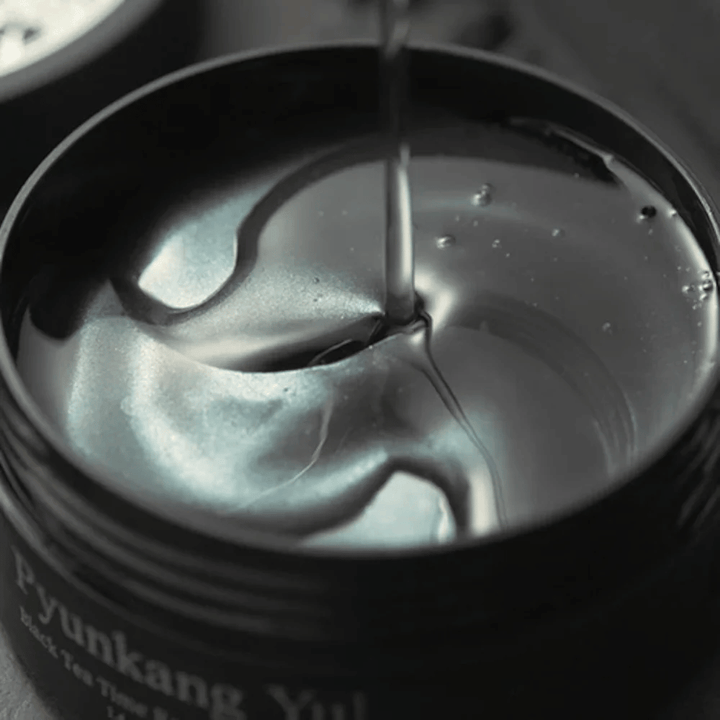 Gros plan sur des patchs contour des yeux en hydrogel noirs dans un pot ouvert de Pyunkang Yul. Les patchs brillants baignent dans le sérum et mettent en valeur le caractère hydratant et luxueux du produit.