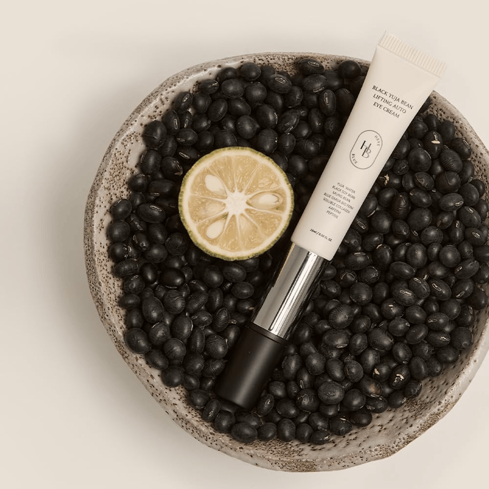 Black Yuja Bean Lifting Auto Eye Cream placé dans un bol rempli de haricots noirs et d'un agrume coupé en deux – une représentation visuelle des ingrédients naturels du produit.