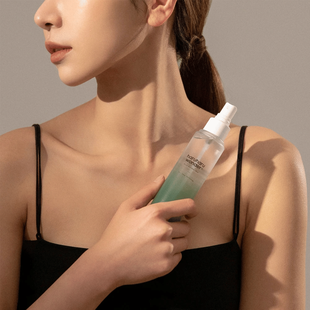 Image de HaruHaru Wonder Black Bamboo Mist tenue par une femme. Le produit est dans un flacon pulvérisateur transparent avec une teinte verte et une pompe blanche. La femme a un look décontracté, porte un haut noir et se concentre sur l'utilisation de la brume pour donner à la peau hydratation et éclat. Le flacon fait partie d'une routine de soins de la peau.