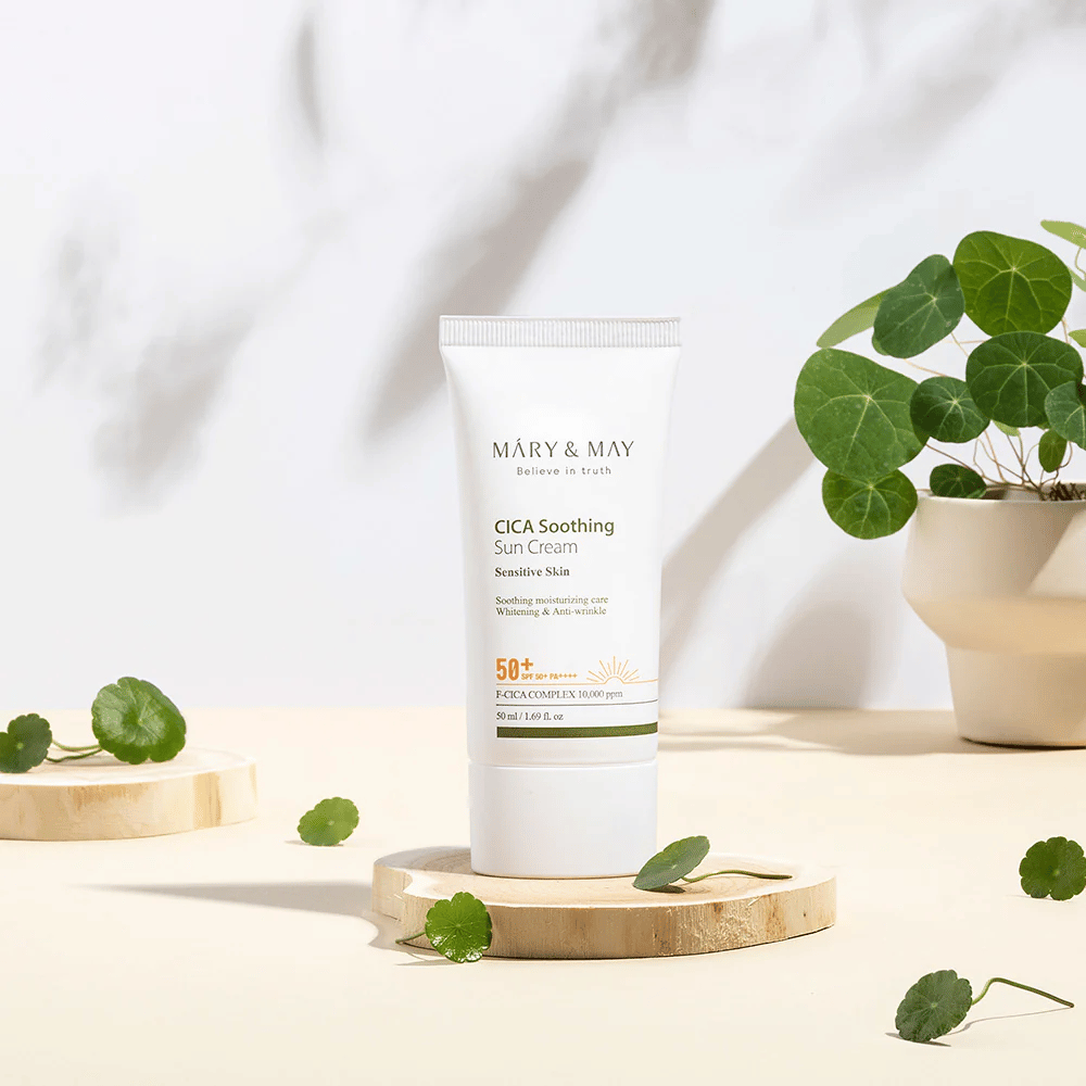 Mary&May CICA Soothing Sun Cream placé sur un podium en bois avec des feuilles de centella vertes tout autour. L’image souligne les ingrédients apaisants pour la peau et l’inspiration naturelle derrière le produit.