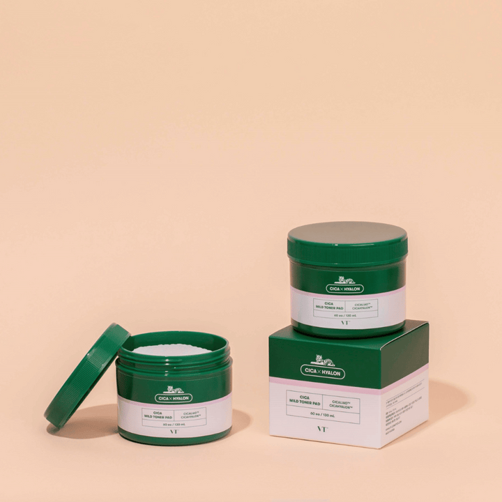 Två gröna burkar med VT Cica Mild Toner Pad, en öppen som visar de vita tonerpadarna och en stängd placerad ovanpå sin produktförpackning, mot beige bakgrund.