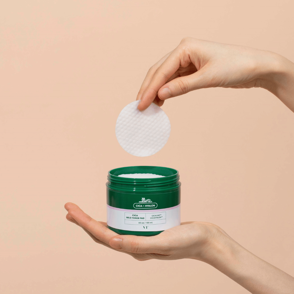Hand som håller i en grön burk med VT Cica Mild Toner Pad och lyfter upp en rund, vit tonerpad över burken mot beige bakgrund.