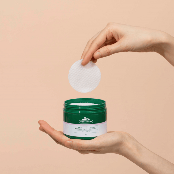 Hand som håller i en grön burk med VT Cica Mild Toner Pad och lyfter upp en rund, vit tonerpad över burken mot beige bakgrund.