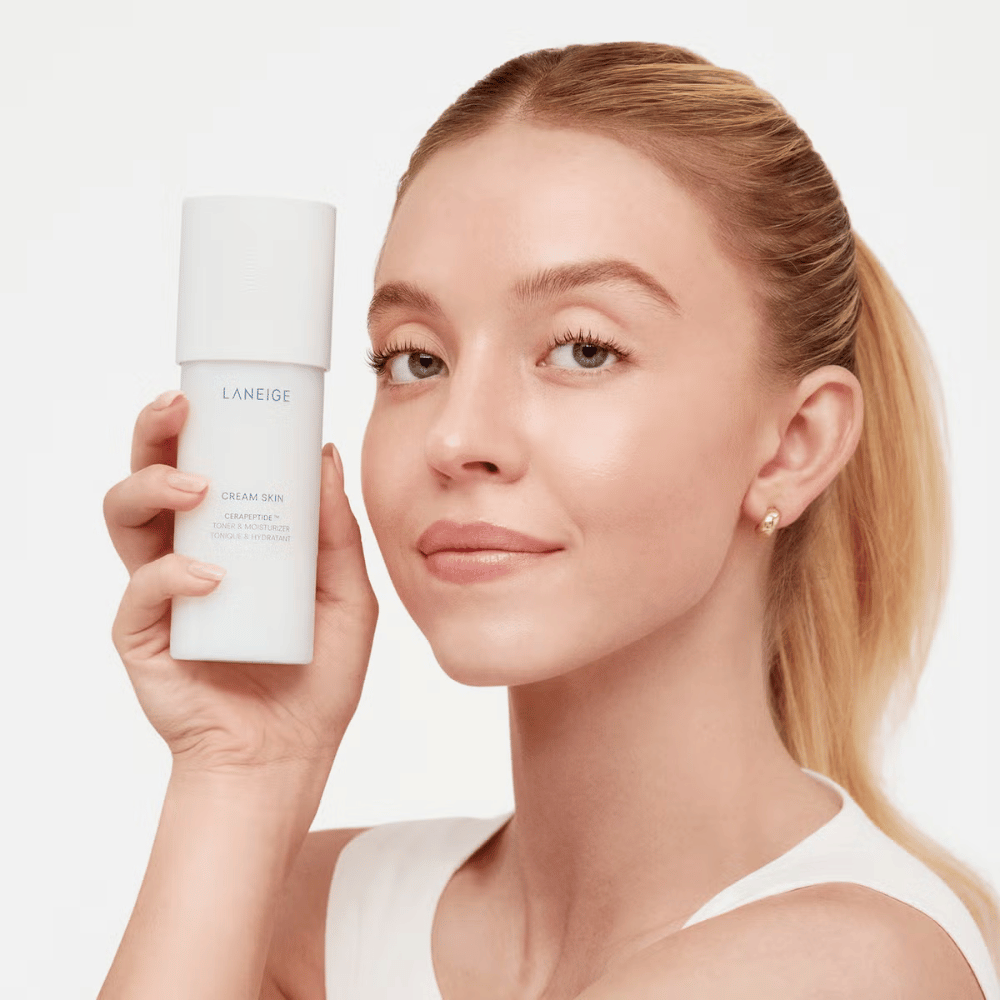 Une personne tient Laneige Cream Skin Cerapeptide Refiner dans une bouteille blanche. La personne sourit et montre le produit, qui est commercialisé comme un toner hydratant et un produit de soin de la peau.