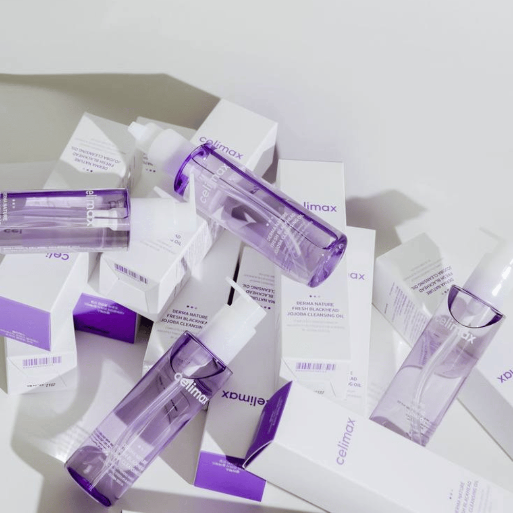 Celimax Derma Nature Fresh Blackhead Jojoba Cleansing Oil dans des bouteilles violettes placées parmi des emballages de produits blancs avec des détails violets.