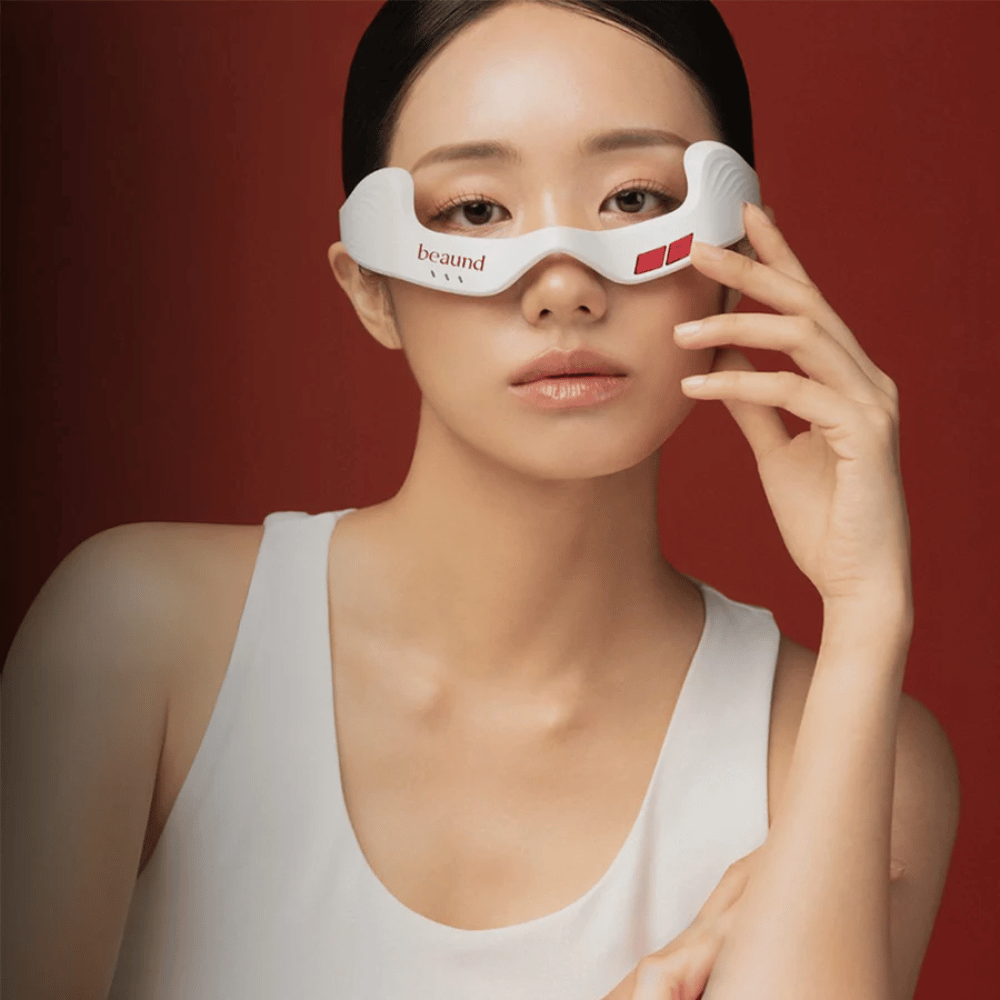 Image de modèle où une personne utilise Beaund Eye Rejuv, un appareil LED futuriste pour la zone autour des yeux, sur un fond rouge élégant.