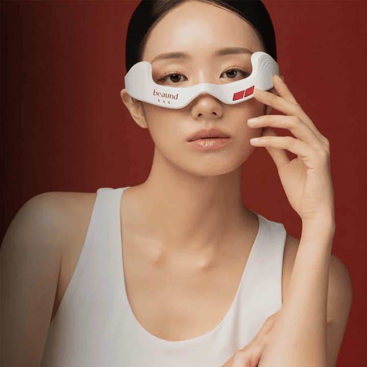 Image de modèle où une personne utilise Beaund Eye Rejuv, un appareil LED futuriste pour la zone autour des yeux, sur un fond rouge élégant.