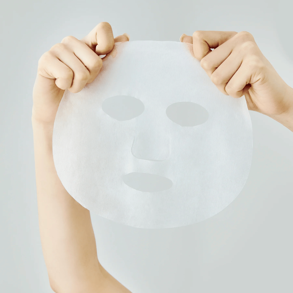 Deux mains tiennent un masque en feuille blanc contre un fond gris clair, avec des découpes visibles pour les yeux, le nez et la bouche.