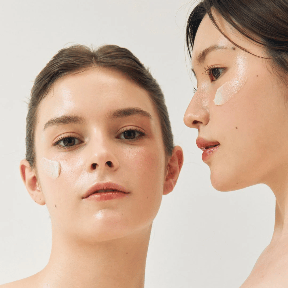 Deux femmes avec des masques faciaux appliqués sur des parties de leurs visages. L'une des femmes a un masque sur la joue et le nez, tandis que l'autre l'a sous l'œil et sur la joue. Elles ont un maquillage naturel et ont l'air fraîches, ce qui met en valeur l'effet hydratant et éclaircissant des masques.