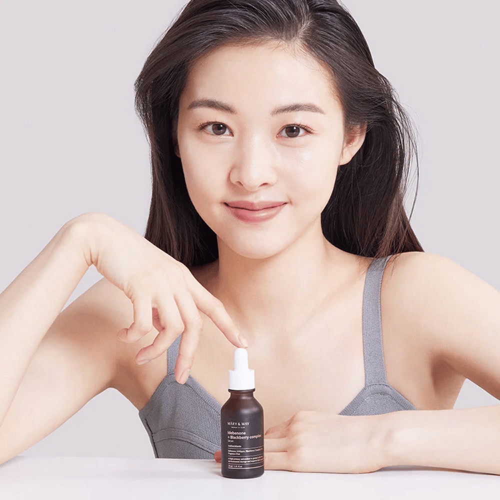Une femme aux longs cheveux foncés tient MARY & MAY Idebenone + Blackberry Complex Serum, une bouteille en verre foncé avec une pipette. Elle a un sourire joyeux et pointe la bouteille avec son autre main. La femme est vêtue d'un haut gris sans manches et semble se trouver dans un environnement lumineux et simple, ce qui met en valeur l'accent du produit sur les soins de la peau et la beauté.