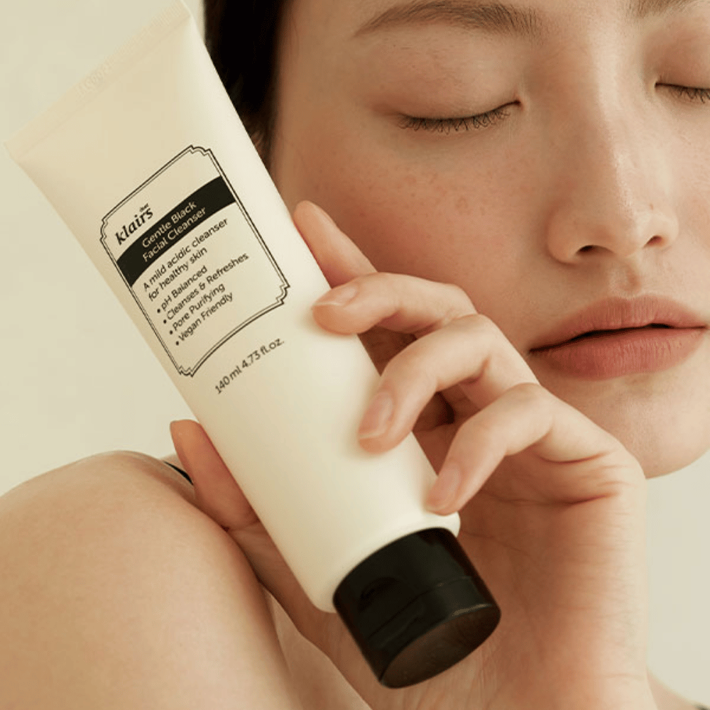 Une personne tient un tube de Klairs Gentle Black Facial Cleanser contre sa joue, les yeux fermés et le teint naturel visible en arrière-plan.