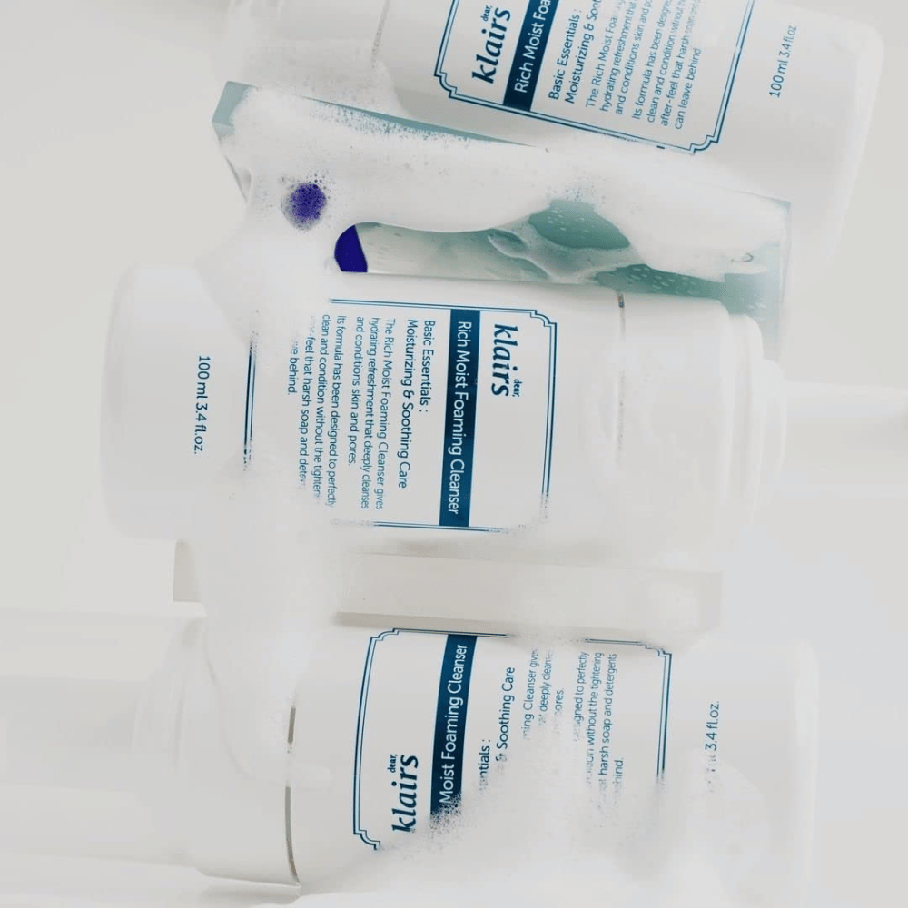 Trois bouteilles de Klairs Rich Moist Foaming Cleanser couvertes de mousse et de gouttes d'eau, placées sur une surface blanche.