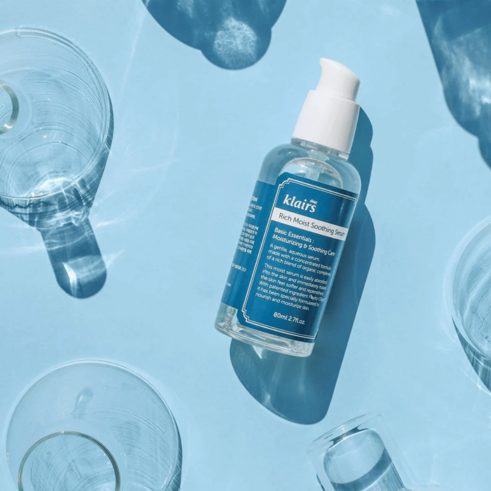 Une bouteille Klairs Rich Moist Soothing Serum parmi des verres d'eau avec des reflets sur un fond bleu.