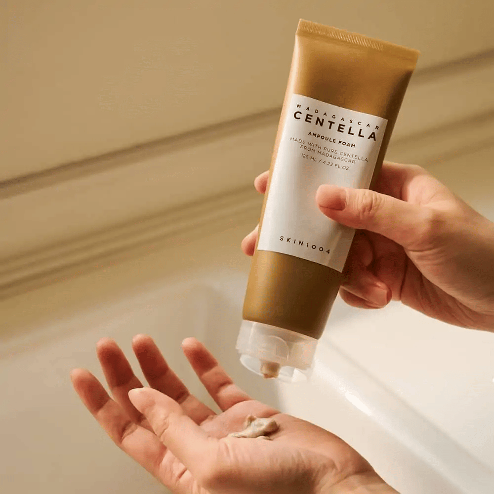 Le produit sur l'image est "Madagascar Centella Ampoule Foam" de SKIN1004. C'est un nettoyant pour le visage sous forme de mousse, fabriqué à partir de Centella de Madagascar. Il contient 125 ml / 4.22 fl.oz. Une main presse une petite quantité de produit pour l'utilisation.