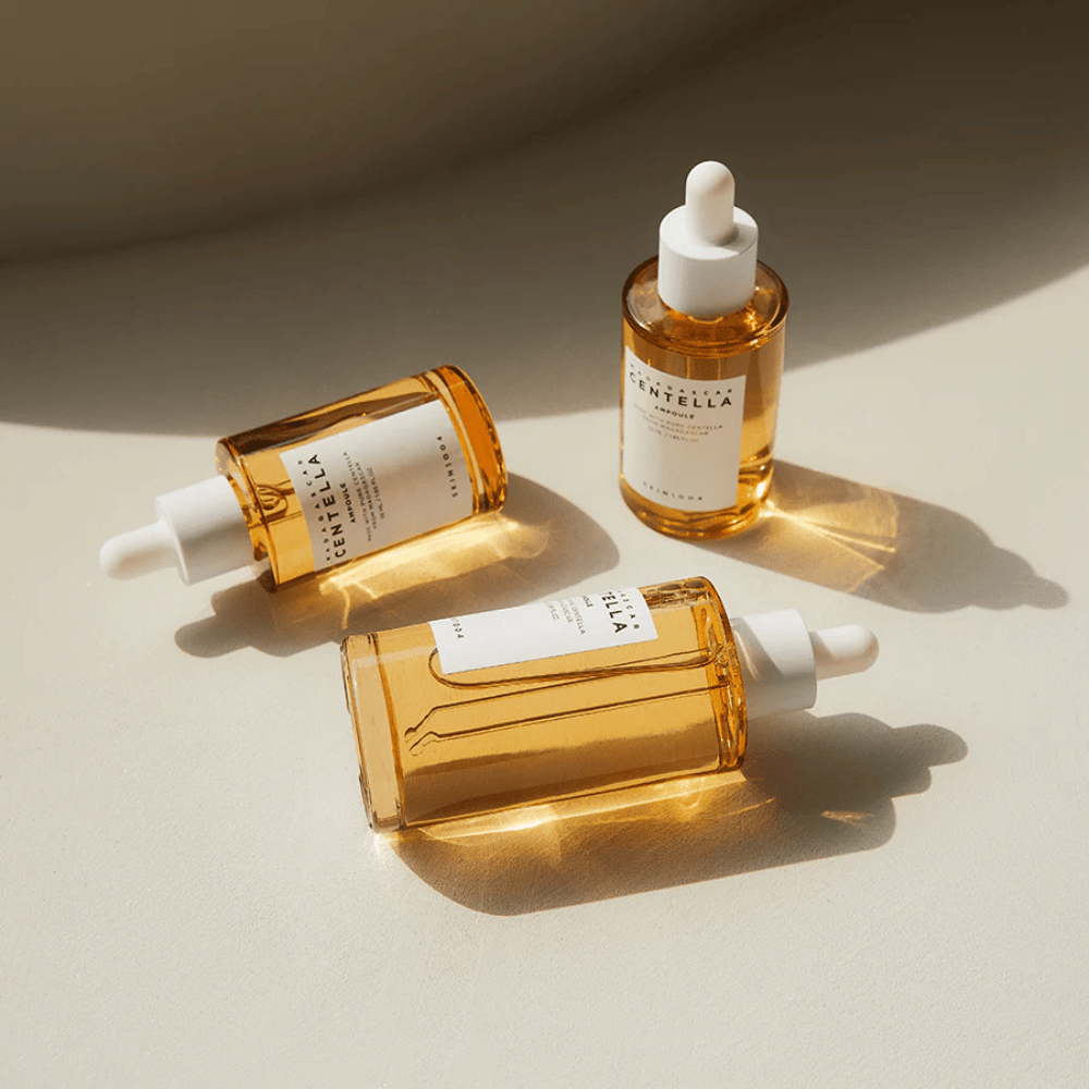 L'image montre trois bouteilles de "Madagascar Centella Asiatica 100 Ampoule". Elles sont placées sur une surface où la lumière du soleil crée une belle réflexion à travers le contenu doré des bouteilles. Le produit semble faire partie d'une série de soins de la peau utilisant la centella asiatica, un ingrédient connu pour ses propriétés apaisantes. Le design des bouteilles est simple et moderne, ce qui suggère qu'il s'agit d'une série de soins de la peau premium.