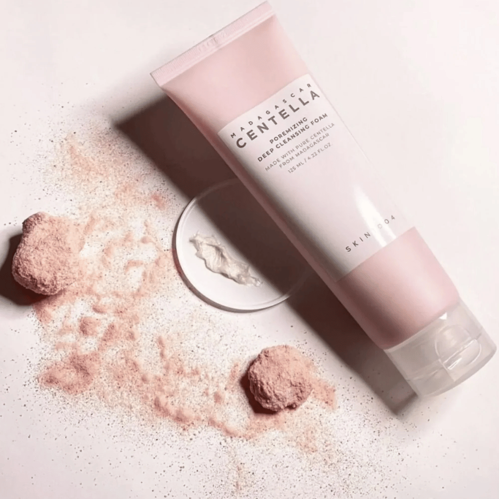 Madagascar Centella Poremizing Deep Cleansing Foam est présenté dans un tube rose. Les tubes sont posés sur une surface claire avec de la poudre rose qui donne une sensation de pureté et de fraîcheur. Le produit est conçu pour nettoyer en profondeur la peau à l'aide de centella pure de Madagascar.