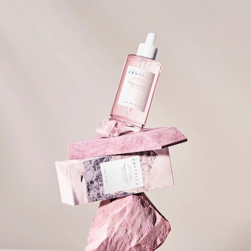 Flacon transparent avec ampoule de Centella rose de SKIN1004, équilibré sur des blocs de pierre, avec un emballage en carton en dessous, sur un fond neutre.