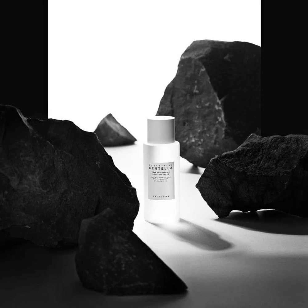 Bouteille blanche SKIN1004 Madagascar Centella Tone Brightening Boosting Toner  placée de manière dramatique parmi de grosses pierres noires sur fond blanc.