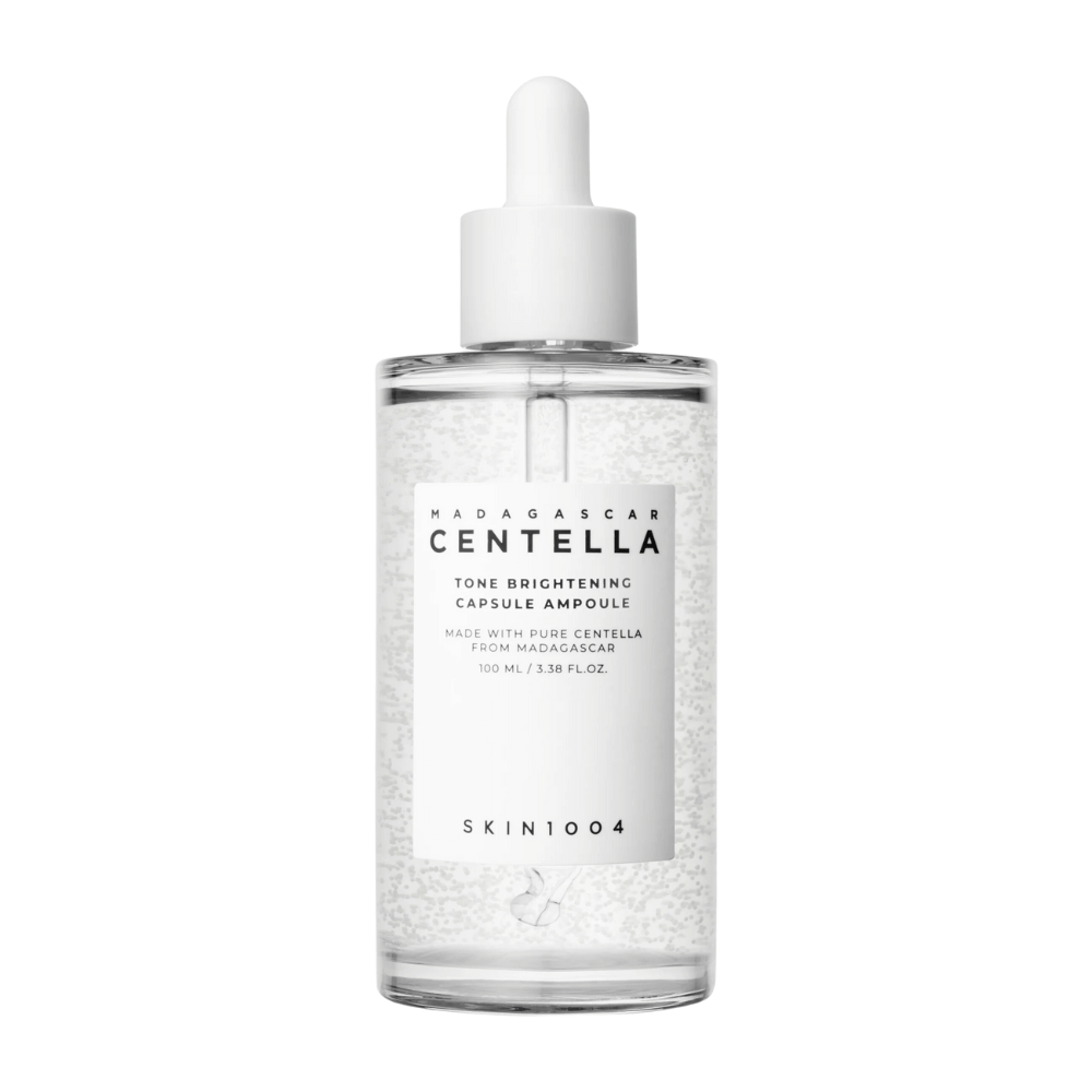 L'image montre une bouteille de "SKIN1004 Madagascar Centella Tone Brightening Capsule Ampoule". C'est une bouteille transparente avec un applicateur compte-gouttes blanc. L'étiquette indique que le produit contient des ingrédients purs de centella de Madagascar. Le contenu de la bouteille semble contenir de petites capsules ou particules, ce qui peut faire partie de ses propriétés éclaircissantes pour la peau. La bouteille contient 100 ml / 3.38 fl.oz. du produit.