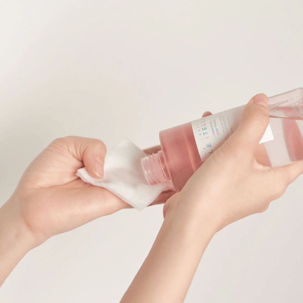Une personne verse Madagascar Centella Poremizing Clear Toner d'une bouteille transparente sur un coton. Le produit, qui a une teinte rose, est utilisé pour rafraîchir et nettoyer la peau. Les mains sont visibles, montrant l'application du toner.