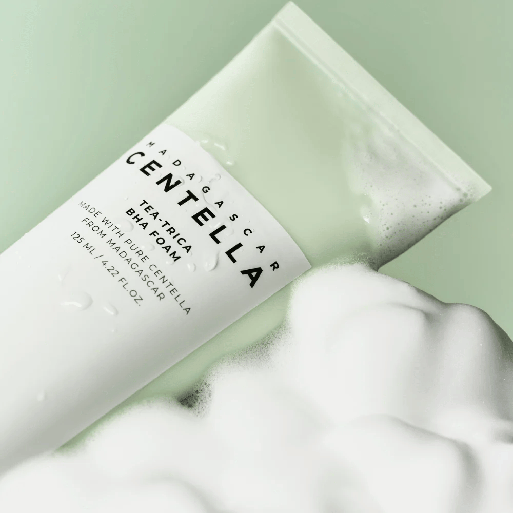 Un produit de type mousse nettoyante, "Madagascar Centella Tea-trica BHA Foam", est partiellement visible avec de la mousse autour. Le produit a un design vert et blanc, et les textes "Madagascar Centella" et "Tea-trica BHA Foam" sont clairement visibles. Le volume est de 125 ml.