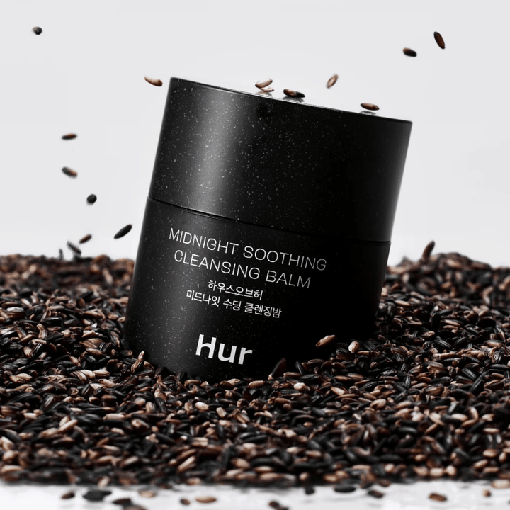 Comment Midnight Soothing Cleansing Balm dans un pot noir placé sur des graines noires. Baume nettoyant apaisant qui dissout le maquillage et les impuretés tout en prenant soin de la peau.