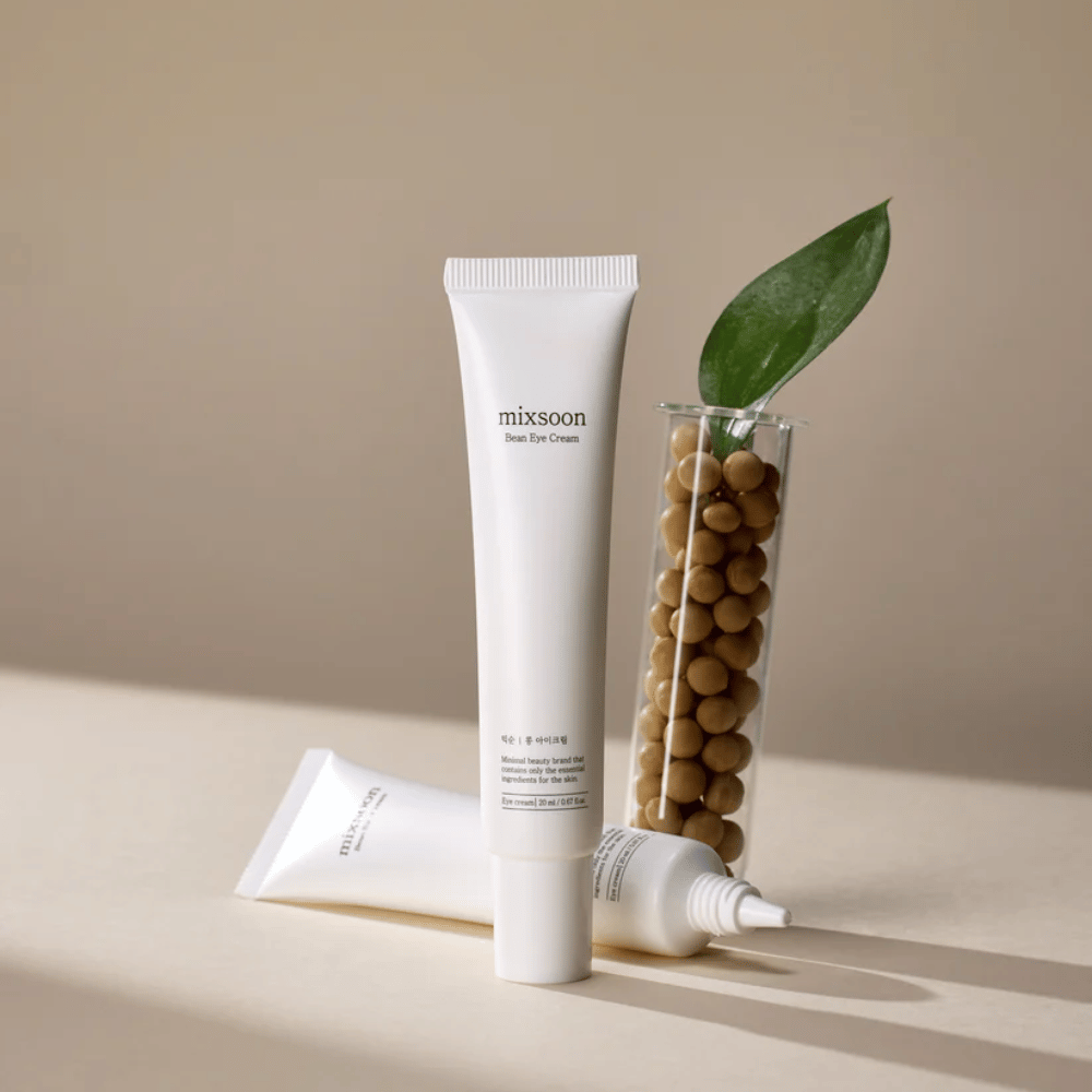 Deux tubes Mixsoon Bean Eye Cream placés à côté d'un tube à essai rempli de graines de soja et d'une feuille verte, sur une surface beige avec des jeux d'ombres.