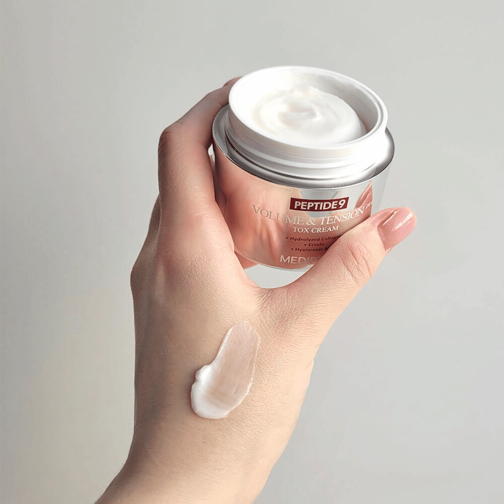 Main tenant MEDI-PEEL Peptide 9 Volume & Tension Tox Cream avec une noisette de crème sur le dos de la main. La formule soyeuse hydrate et donne à la peau fermeté et élasticité.