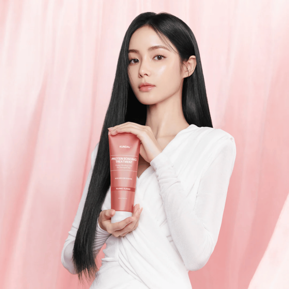 Une femme avec de longs cheveux brillants tient KUNDAL Protein Bonding Treatment Violet Muguet sur un fond rose.