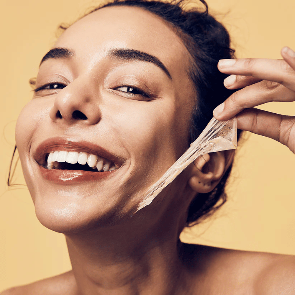 L'image montre une femme avec un grand sourire en train de retirer un masque peel-off de son visage. La femme a l'air heureuse et satisfaite tout en utilisant le produit de beauté, ce qui transmet un sentiment de bien-être et de soin de la peau.