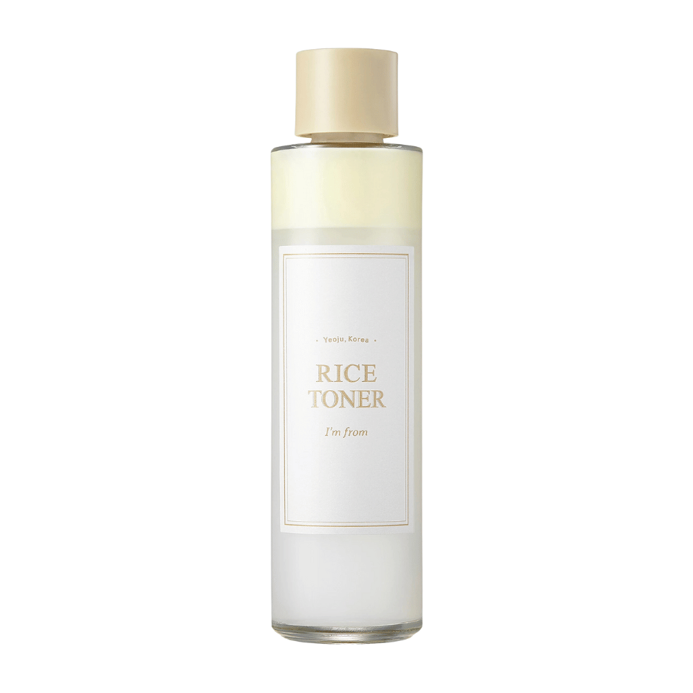 Une bouteille transparente avec un bouchon doré et une étiquette indiquant "Rice Toner I'm from Yeojü, Corée". La bouteille contient un liquide clair, probablement une lotion tonique à base d'extrait de riz. Le design est épuré et minimaliste. Le fond est blanc.
