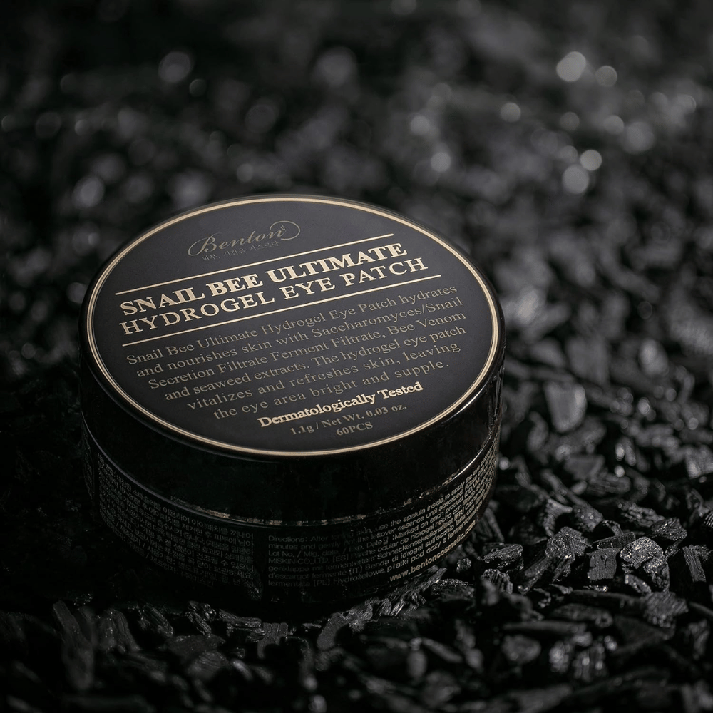 En svart rund burk med Benton Snail Bee Ultimate Hydrogel Eye Patch placerad på en mörk yta. Burkens etikett har guldtext som beskriver produktens innehåll och fördelar. Den innehåller 60 ögonlappar.