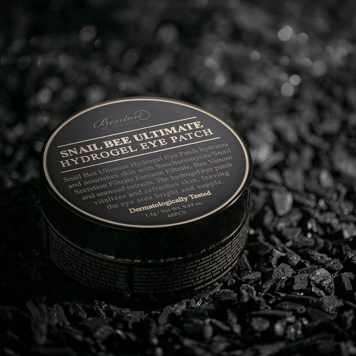 En svart rund burk med Benton Snail Bee Ultimate Hydrogel Eye Patch placerad på en mörk yta. Burkens etikett har guldtext som beskriver produktens innehåll och fördelar. Den innehåller 60 ögonlappar.