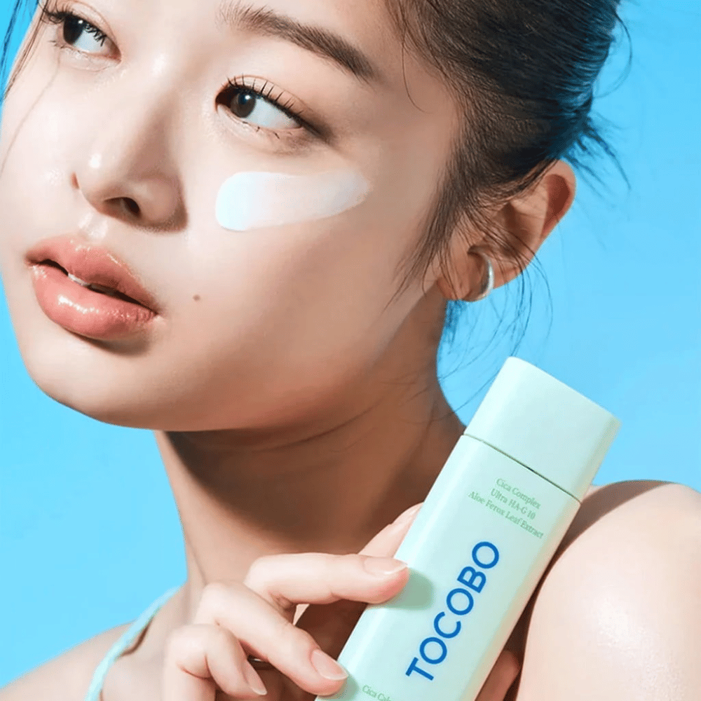 Mannequin avec crème solaire sous l'œil tenant TOCOBO Cica Calming Sun Serum dans un emballage vert menthe sur fond bleu.
