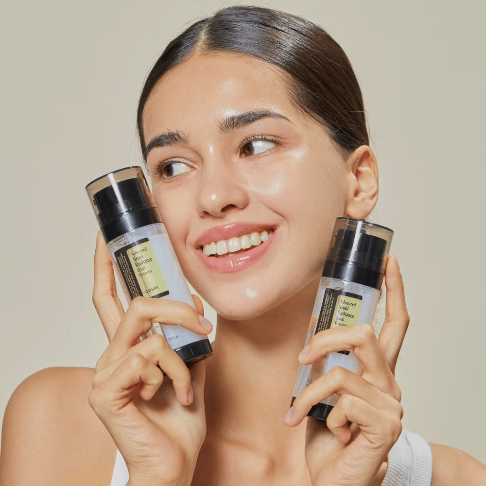 Femme souriante avec peau éclatante tenant deux flacons COSRX Advanced Snail Radiance Dual Essence 80 ml près du visage sur fond clair.