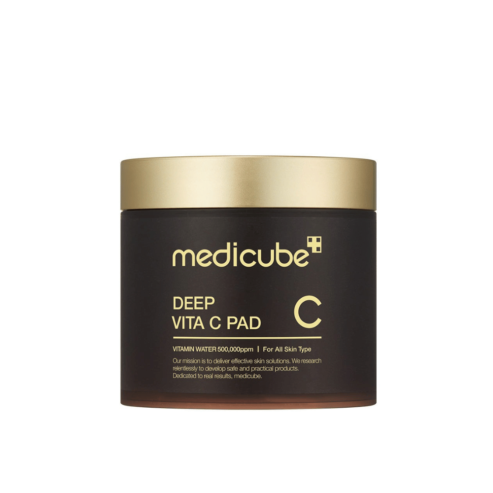 Un pot de medicube, marqué "Deep Vita C Pad", avec un couvercle doré et un corps sombre. Le texte sur le pot indique de la vitamine C et une adéquation pour tous les types de peau. Le fond est neutre.