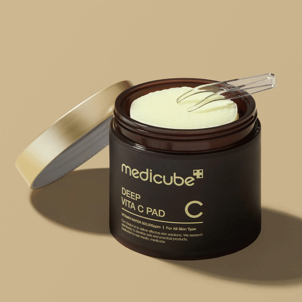 Un pot avec medicube Deep Vita C Pad. Le pot est brun foncé avec du texte doré et une étiquette blanche inversée avec des informations sur la vitamine C. L'intérieur du couvercle est doré, et une pince transparente repose sur les tampons de coton jaune clair à l'intérieur. Le fond est beige uni.
